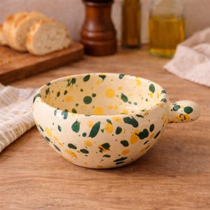 Op de afbeelding: Crèmekleurige keramische soepkom met een handvat, besprenkeld met gele en groene verf. De kom staat op een houten oppervlak, met brood en een houten plank op de achtergrond. Een gestreepte doek is links.