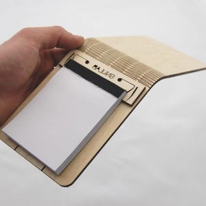 Puede incluir: Un portapapeles de madera con un bloc de notas blanco y encuadernación negra. El bloc de notas se sujeta dentro de un marco de madera cortado con láser. La palabra "saline" está grabada en la madera. El bloc de notas está abierto, revelando las páginas en blanco.