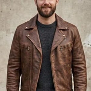K&ouml;nnte beinhalten: Braune Lederjacke im klassischen Biker-Stil. Die Jacke hat einen Reverskragen, einen asymmetrischen Rei&szlig;verschluss und mehrere Taschen. Das Model tr&auml;gt einen dunkelgrauen Pullover darunter. Die Jacke scheint aus hochwertigem Leder zu sein.