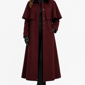 May include: A floor-length, deep burgundy wool-blend coat featuring a structured cape overlay, a dark velvet collar, and a front button closure. The garment is worn over a black turtleneck, black trousers, black leather gloves, and black boots.