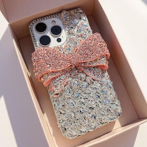 Pode incluir: Uma capa de telemóvel adornada com um grande laço rosa. A capa é coberta por cristais e strass brilhantes, com um esquema de cores prateado e transparente. O laço é num tom de rosa mais claro e adiciona um toque feminino. A capa do telemóvel está numa caixa rosa.