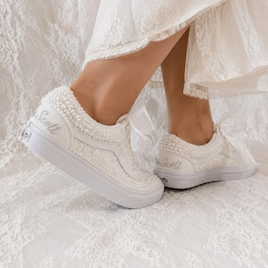 May include: A pair of white low-top trainers covered in small white pearls, featuring white ribbon laces and the text "Mrs. Scott" embroidered in silver on the heel. The trainers have a white rubber sole and are shown against a white lace fabric.