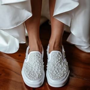 Puede incluir: Zapatillas blancas sin cordones adornadas con numerosas perlas blancas pequeñas y redondas. Los zapatos tienen una suela de goma blanca y están sobre un suelo de madera. Las zapatillas son de estilo nupcial o de ocasión especial.