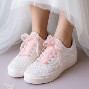 Könnte beinhalten: Weiße Sneaker, bedeckt mit kleinen, runden, perlenartigen Verzierungen. Die Sneaker haben rosa Schnürsenkel und große rosa Schleifen oben. Die Sohlen sind weiß, und die Schuhe scheinen Teil eines Brautensembles zu sein.