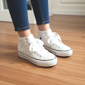 May include: White high-top sneakers embellished with small, round, pearl-like beads. The shoes have white laces, a black star logo, and a black stripe along the white sole. The sneakers are worn with blue jeans.