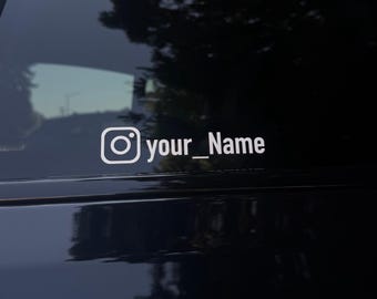 May include: White Instagram logo decal with the text "your_Name" on a dark surface, likely a car window. The decal is clean and simple, with a modern aesthetic. The background is blurred, suggesting an outdoor setting.