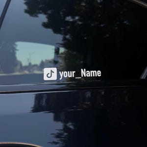 Könnte beinhalten: Weißer Vinyl-Aufkleber mit dem TikTok-Logo und dem Text "your_Name". Der Aufkleber ist auf der Scheibe eines dunkel gefärbten Fahrzeugs angebracht. Der Hintergrund zeigt eine verschwommene Ansicht von Bäumen und einer Straße.