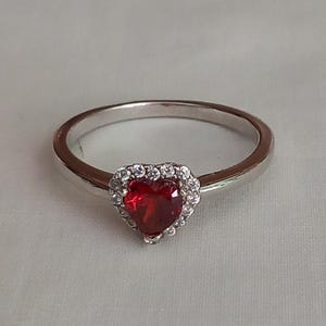 Peut inclure: Une bague en argent avec une pierre rouge en forme de cœur entourée d'un halo de petites pierres claires.