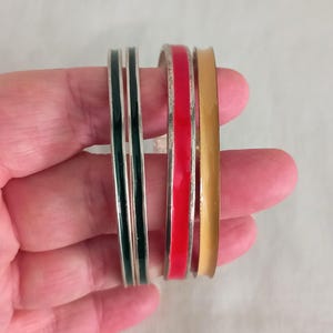 Puede incluir: Cuatro pulseras de plata con esmalte de color. Las pulseras están apiladas una encima de la otra. La pulsera superior es roja, la segunda es dorada, la tercera es plateada y la pulsera inferior es verde.