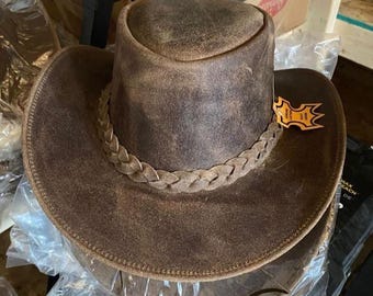 May include: A brown leather cowboy hat with a braided band around the crown. The hat has a wide brim and a distressed finish. A small tag is attached to the brim. The hat is likely new and in its packaging.