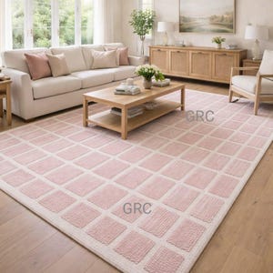 Peut inclure: Un grand tapis rectangulaire avec un motif géométrique dans un salon. Le tapis présente un motif répétitif de carrés rose clair soulignés de blanc. La pièce comprend un canapé blanc, une table basse en bois et de la lumière naturelle.