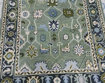 Könnte beinhalten: Ein rechteckiger, salbeigrüner Teppich mit einem komplizierten floralen und geometrischen Muster. Der Teppich hat ein zentrales Feld mit sich wiederholenden floralen Motiven in Blau-, Weiß- und Grüntönen, das von einem dunkelblauen Rand mit zusätzlichen geometrischen Designs umrahmt wird. Der Teppich hat einen Fransen.