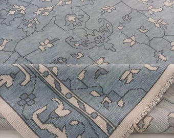 May include: A light blue rug with a floral pattern and a cream-colored border. The rug features a detailed design with various floral and geometric shapes, creating a classic and elegant look. The rug is partially rolled up, revealing its fringed edge.