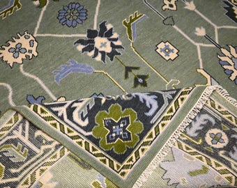 Könnte beinhalten: Ein salbeigrüner Teppich mit einem detaillierten Blumenmuster in Blau-, Creme- und Grüntönen. Das Design umfasst stilisierte Blumen und geometrische Formen. Eine gefaltete Ecke zeigt den gefransten Rand des Teppichs.