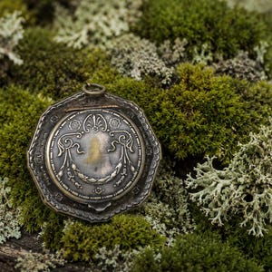 Puede incluir: Un relicario de metal plateado con forma octogonal y patrones de volutas en relieve. El relicario presenta un diseño circular central con motivos florales y un pequeño lazo de metal en la parte superior para sujetarlo a una cadena.