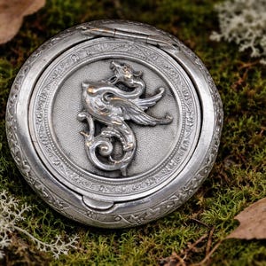 May include: A round, silver-colored compact with a detailed dragon relief on the lid. The dragon is centered within a circular frame, surrounded by ornate patterns. The compact is set against a backdrop of green moss and fallen leaves.
