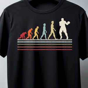 May include: Black t-shirt featuring a retro-style evolution graphic. The graphic shows a series of figures evolving from ape to boxer, with colorful horizontal stripes below. The design is printed on a black crew neck t-shirt.