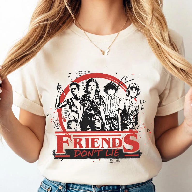 May include: Cream-colored t-shirt featuring a graphic of four characters from the show "Stranger Things" inside a red circle. The text "FRIENDS DON'T LIE" is printed in red and black below the graphic.
