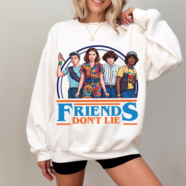 May include: White sweatshirt featuring a graphic of four characters from the show "Stranger Things" inside a blue circle. The text "FRIENDS DON'T LIE" is printed in orange and blue below the graphic. The characters are wearing colorful outfits.