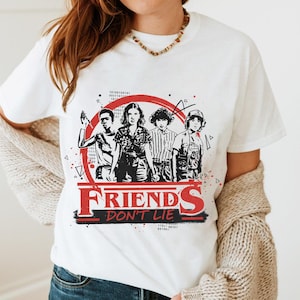 May include: White t-shirt featuring a graphic of four characters inside a red circle, with the words "FRIENDS DON'T LIE" in red and black. The design is a reference to a popular science fiction series.