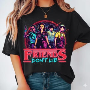 May include: Black t-shirt featuring a graphic of four characters from the show "Stranger Things" with the text "FRIENDS DON'T LIE" in red and blue. The design is set against a red circular background with a starry effect.