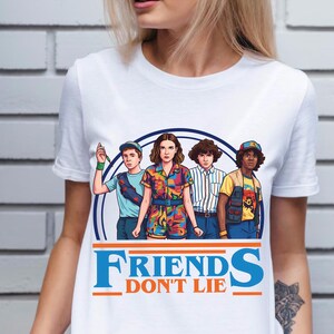 May include: White t-shirt featuring a graphic of four characters from the show "Stranger Things" inside a blue circle. The text "FRIENDS DON'T LIE" is printed in blue and orange below the graphic. The characters are in colorful outfits.
