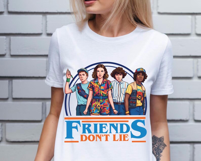 May include: White t-shirt featuring a graphic of four characters from the show "Stranger Things" inside a blue circle. The text "FRIENDS DON'T LIE" is printed in blue and orange below the graphic. The characters are in colorful outfits.