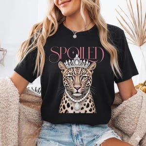 Op de afbeelding: Zwart T-shirt met een luipaardillustratie met een kroon en een parelketting. Het woord "SPOILED" staat in roze letters boven de luipaard gedrukt. De luipaard heeft een gevlekte vacht en een koninklijke uitdrukking.
