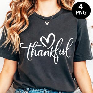 May include: Dark grey t-shirt with the word "thankful" in white script, a heart above the word. The shirt is a crew neck and has short sleeves. A small butterfly pendant necklace is visible.