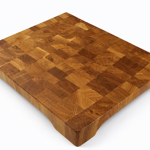 May include: A square, end-grain butcher block cutting board made of light and dark brown wood. The board has a mosaic-like pattern and is raised slightly off the surface with small feet. This durable board is ideal for food preparation.