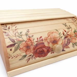 May include: Wooden bread box with a light wood finish and a floral design. The design features large, watercolor-style roses and foliage in shades of red, orange, and brown. The box has a curved front and a wooden knob.