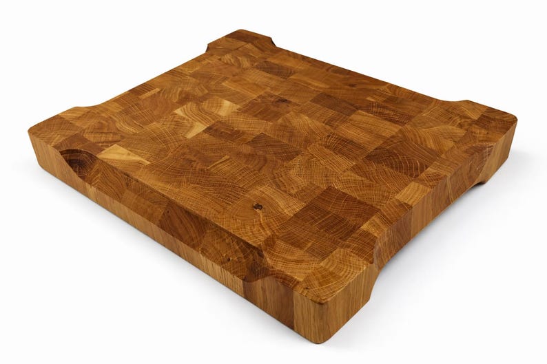 May include: A square, end-grain butcher block cutting board made of light brown wood. The board has a mosaic-like pattern and rounded corners with finger grips. The wood grain is visible throughout the board, which is ideal for kitchen use.