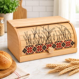 May include: A light-colored wooden bread box with a roll-top lid featuring a black ink illustration of wheat stalks and a horizontal band of red and black geometric star patterns. A small round wooden knob is attached to the front of the lid.