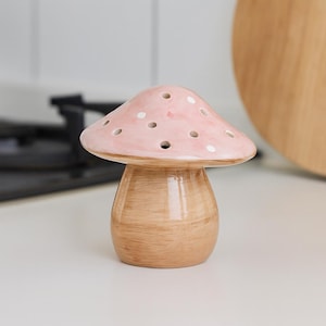 Op de afbeelding: Een keramisch paddenstoelvormig object met een lichtroze kap met witte stippen en een lichtbruine steel. De paddenstoel heeft verschillende gaten op de kap. Het object is waarschijnlijk een decoratief item of een keukenaccessoire.
