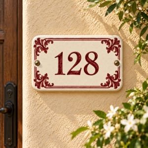 Peut inclure: Une plaque de numéro de maison rectangulaire en céramique affichant les chiffres 128 en rouge foncé. La plaque présente un fond crème avec une bordure rouge foncé et des motifs de volutes décoratives dans chaque coin, fixée par deux vis.
