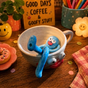 May include: A light blue ceramic mug featuring a sculpted blue character with white eyes and red lips sitting inside. The mug itself has a matching face with large white eyes and a red mouth on the exterior, with a handle on the right side.