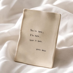 Pode incluir: Uma bandeja retangular de cerâmica na cor off-white com bordas irregulares moldadas à mão. A superfície apresenta um texto manuscrito em preto que diz: You're here, I'm here, love is here. XOXO Kico. A bandeja tem um acabamento fosco e liso.