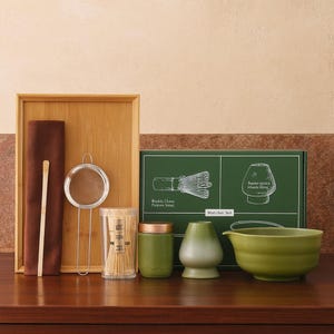 May include: A matcha tea set featuring a wooden tray, whisk, strainer, bamboo scoop, and a green ceramic bowl. The set includes a container of bamboo whisks, a green container, and a green and white ceramic tea cup. A green box with illustrations of a whisk and tea cup is also included.