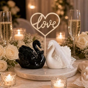 May include: A black swan figurine wearing a small black bow tie and a white swan figurine wearing a white lace veil, positioned together on a circular base. A wooden heart-shaped topper with the word love in cursive script stands between them.