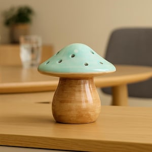 Op de afbeelding: Een keramisch decoratief item in de vorm van een paddenstoel. De paddenstoelkap is lichtblauw met kleine gaatjes en de steel is warmbruin. Het item staat op een houten oppervlak.