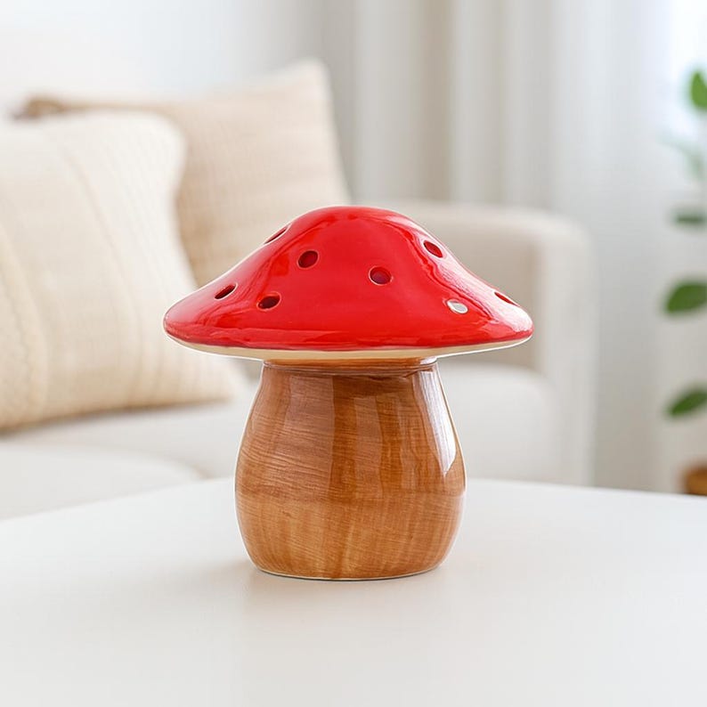 Op de afbeelding: Een keramisch decoratief item in de vorm van een paddenstoel. De felrode hoed heeft verschillende kleine gaatjes. De steel is lichtbruin met een houtnerf. Het item staat op een wit oppervlak.