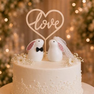 May include: A white cake topper featuring two rabbit figurines, one wearing a black bow tie and the other a white lace veil, positioned beneath a heart-shaped cutout with the word love written in cursive script, all resting on a white cake.