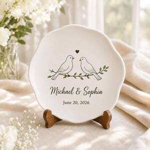 Puede incluir: Un plato de cerámica blanca con borde festoneado que presenta un gráfico de dos pájaros posados en una rama con un pequeño corazón encima. El texto Michael & Sophia y la fecha June 20, 2026 están impresos en negro en el centro del plato.