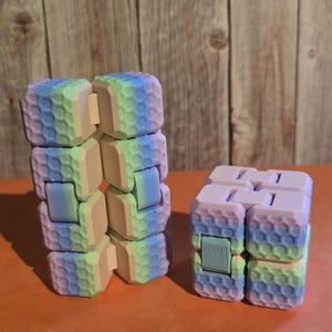 May include: Two multi-coloured fidget cubes. The cubes are made of connected blocks with a honeycomb pattern. The colours include blue, green, pink, and yellow. One cube is in an extended position, and the other is in a folded position.