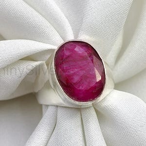 May include: A silver ring featuring a large, oval-shaped, faceted deep pink gemstone set in a smooth metal bezel. The stone displays a textured, opaque surface with natural inclusions, securely mounted on a polished silver band.