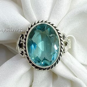May include: A silver ring with an oval, light blue gemstone. The gemstone is surrounded by a twisted rope-like border and ornate silver detailing. The ring is set against a white fabric background. The text "ShinySilverK" is visible.