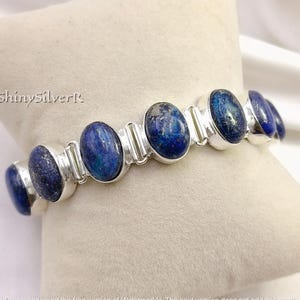 May include: A silver bracelet with oval, deep blue lapis lazuli stones. The stones are set in individual silver bezels and linked together. The bracelet is shown on a light-coloured surface.