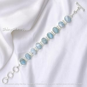 Op de afbeelding: Een zilveren armband met ovale, lichtblauwe edelstenen. De edelstenen zijn gezet in een zilveren band en verbonden door een zilveren ketting. De armband heeft een tuimelsluiting. De tekst "ShinysilverR" is zichtbaar.