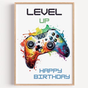 May include: A framed print featuring a colorful watercolor-style video game controller. The controller is splashed with red, orange, yellow, green, and blue paint. Text includes "LEVEL UP," "100," and "HAPPY BIRTHDAY."
