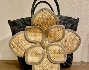 May include: Black woven tote bag with braided handles. A large, decorative flower made of woven straw with a gray border is in front of the bag. The flower has five petals and a circular center, creating a natural, summery aesthetic.